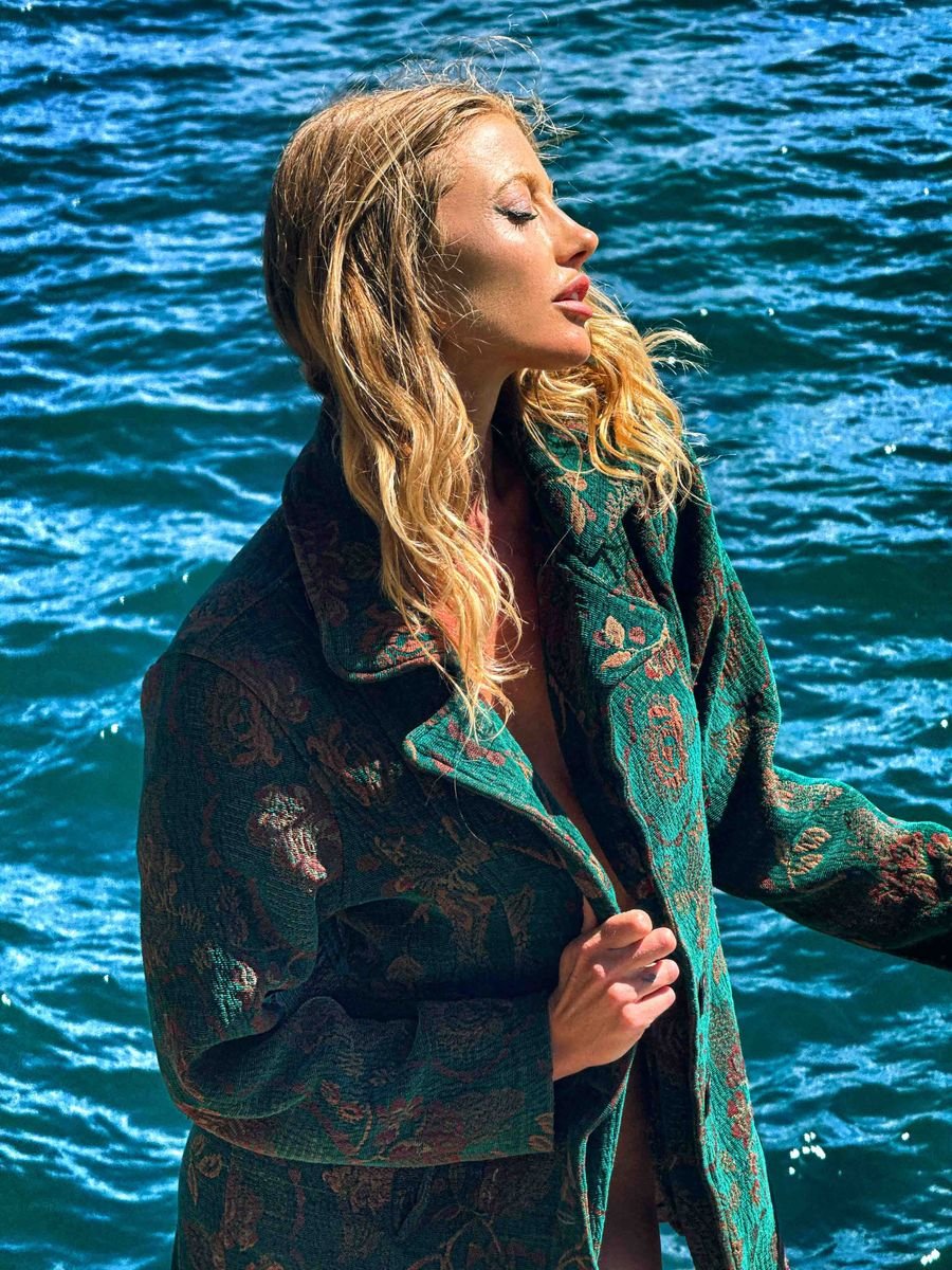 Model in an embroidered green coat in front of deep blue water