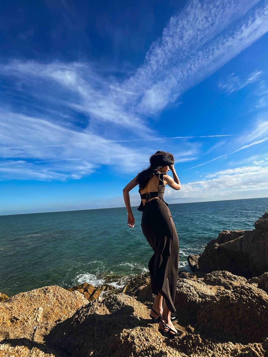 Model in a brown dress on coastal rocks against the sea and a bright sky
