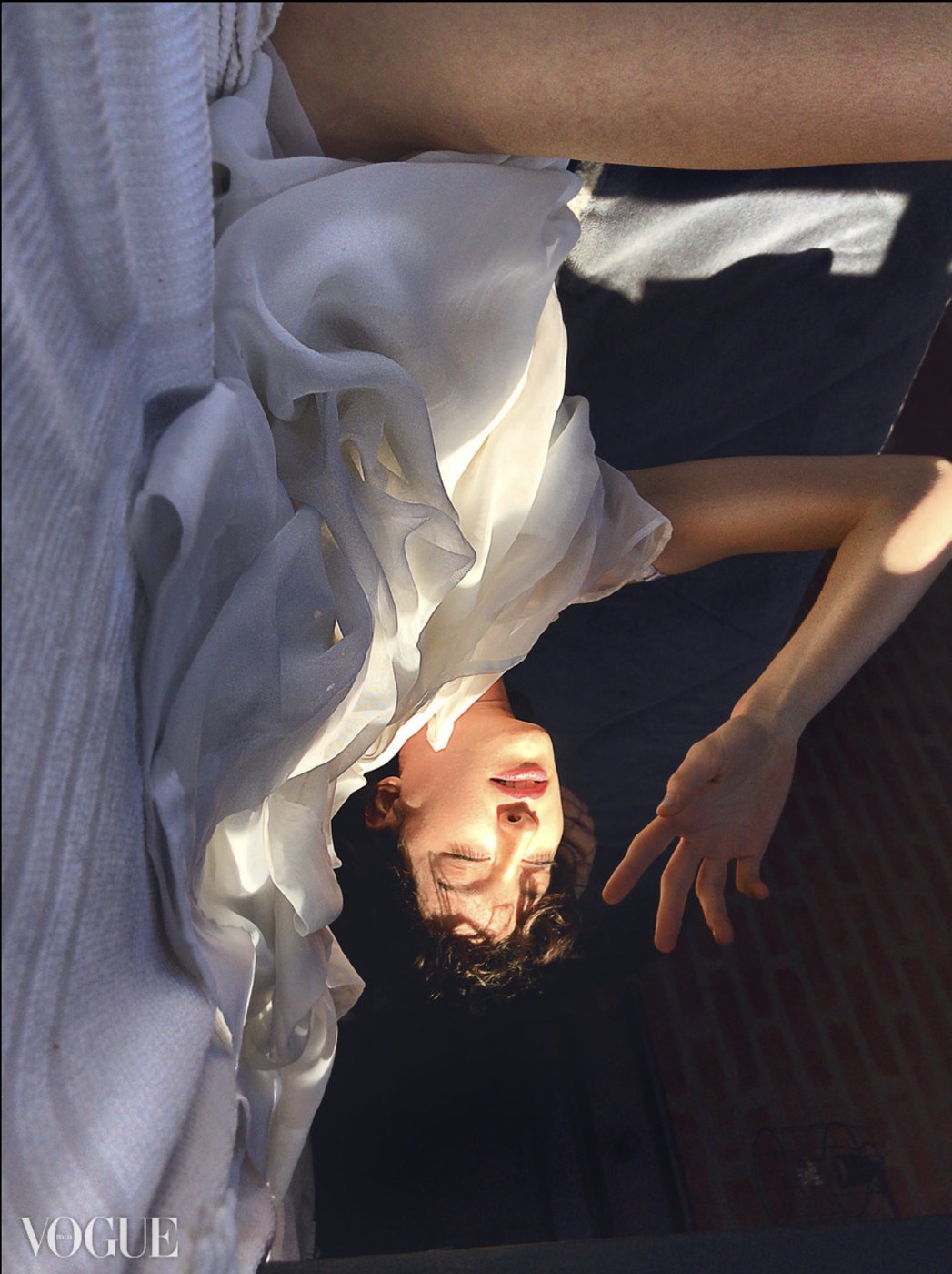 Model in a white chiffon dress photographed from above, eyes closed, hand raised against the light. Published on Photo Vogue